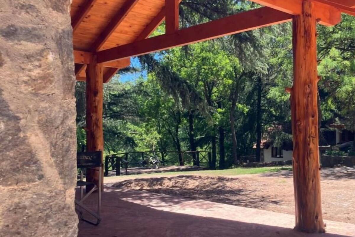 A wooden pavilion with a tiled roof, surrounded by trees and a dirt ground.