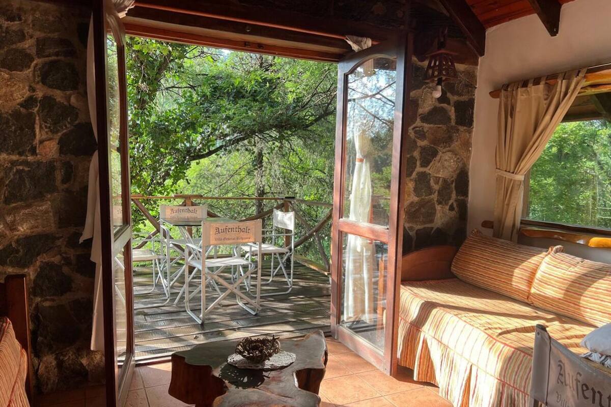 A cozy room with a stone wall, a bed with striped bedding, a table with a decorative item, and a view of an outdoor seating area with chairs and a sign.
