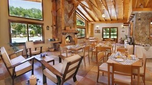 A spacious dining area with wooden tables and chairs, a stone fireplace, and large windows offering a view of the outdoors. - Aufenthalt Cabañas (Villa General Belgrano)
