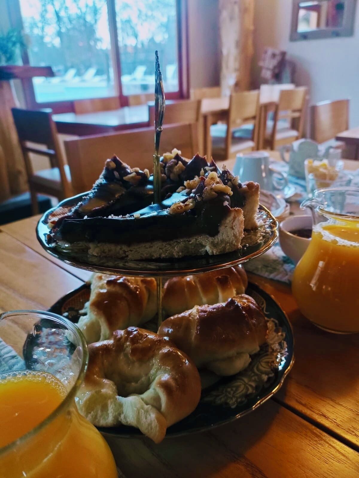Een nagerechtenbord met een plakje taart belegd met noten en een mes, een bord met croissants, en een glas sinaasappelsap.