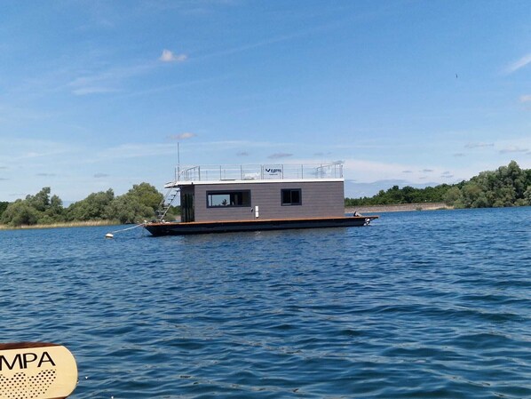 Ein modernes Hausboot mit flachem Dach, einer kleinen Terrasse und einem überdachten Bereich mit Geländer.