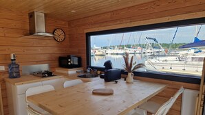Una cabaña de madera con zona de cocina, sillas blancas, una mesa de madera, un reloj y vistas a barcos a través de un ventanal grande.