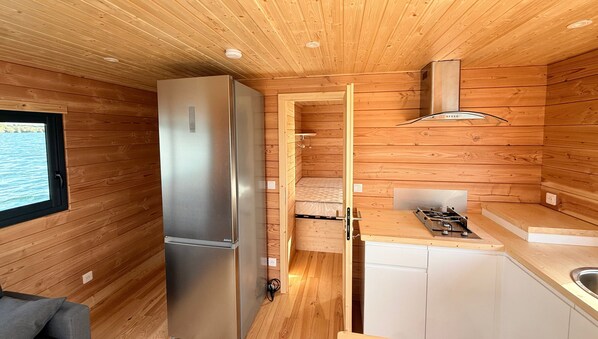 Cocina compacta con paredes de madera, un refrigerador de acero inoxidable, estufa a gas y una ventana pequeña con vista al agua.