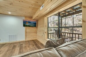 A room with wooden walls and ceiling, a large window with a view of trees, a mounted television, and a sofa. - Renovated Barn with Creek and Waterfall (Cosby)
