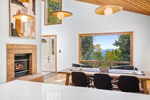 A modern dining area with a fireplace, large windows offering a scenic view, and contemporary lighting fixtures. - Nestled Inn - Trails, Views, Hot Tub, Skiing! (Evergreen)