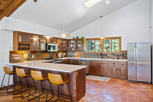 A modern kitchen with a large island, bar stools, wooden cabinets, and a stainless steel refrigerator. - Nestled Inn - Trails, Views, Hot Tub, Skiing! (Evergreen)