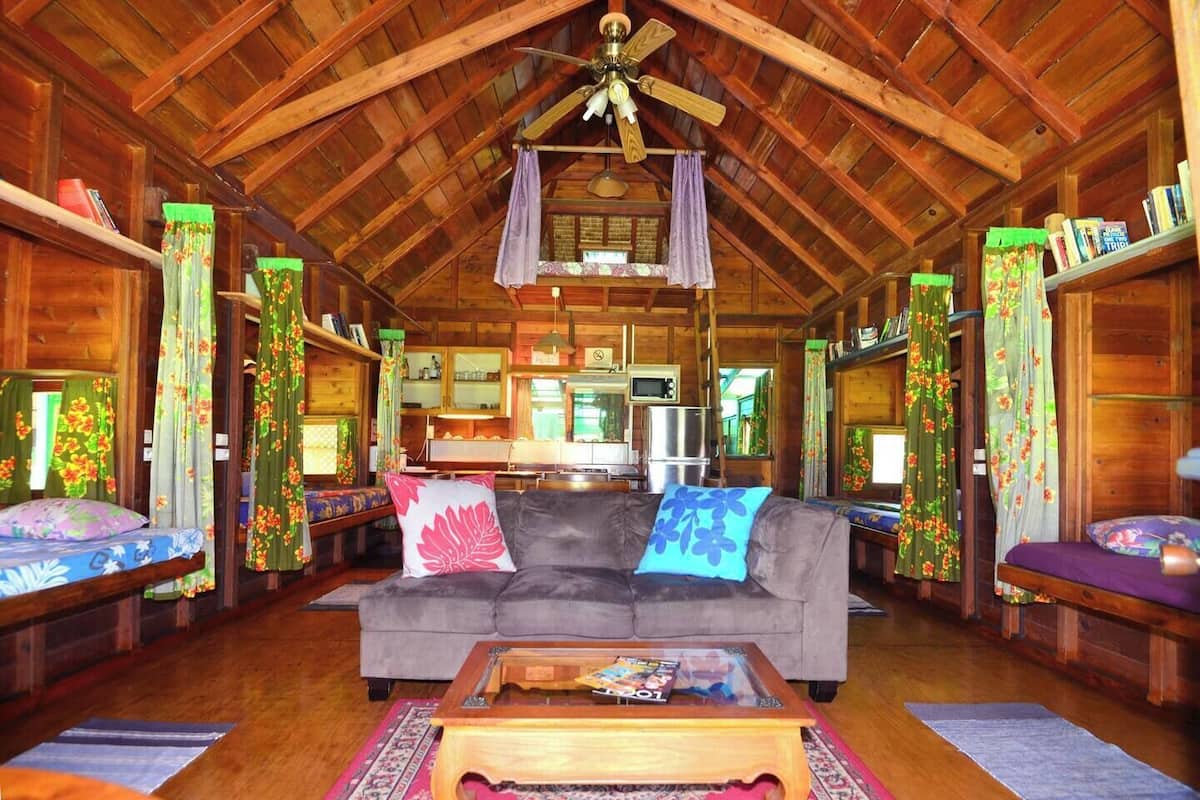 A cozy cabin interior with a wooden ceiling, a sofa with floral cushions, a coffee table, and a kitchen area in the background.