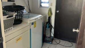 A compact kitchen with a washing machine, a small window with decorative blinds, and a door. - Casa Madeira (Mexicali)