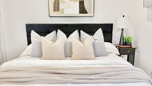A neatly made bed with a white comforter and multiple pillows, a framed abstract artwork above the bed, and a bedside table with a lamp and a small plant. - Modern 2BR Retreat - Fast WiFi, AC & Prime Houston Location (Houston)