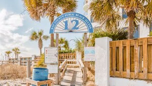 Μια πινακίδα που υποδεικνύει την πρόσβαση στην παραλία της δημόσιας παραλίας του κόλπου Μπέι, με ένα ξύλινο μονοπάτι που οδηγεί στην παραλία, φοίνικες και έναν ξύλινο φράχτη.