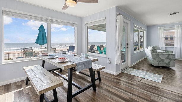 Una habitación con una mesa de madera, banco y un ventilador de techo. Amplias ventanas con vistas a la playa.