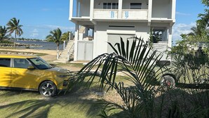 A two-story house with a garage, a yellow car parked in front, and a view of the ocean. - Bay Front Eco-Cottage/2 units/Corozal with WiFi, AC (Corozal)
