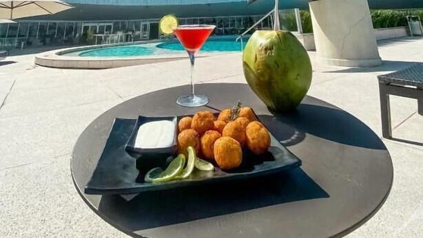 A dining table with a martini, a coconut drink, and fried plantains, set against a backdrop of a tall building and a sculpture, with a pool and umbrellas in the background. - Hotel Nacional (Rio de Janeiro)