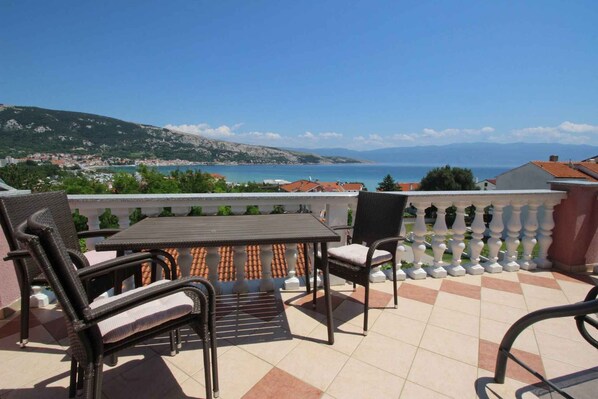 A balcony with a table and chairs, offering a view of a coastal town and the sea.