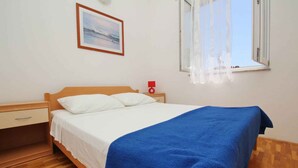 Ein Schlafzimmer mit einem hölzernen Bett, weißer Bettwäsche, einer blauen Decke, einem Nachttisch mit einer roten Lampe, einem gerahmten Bild an der Wand und einem Fenster mit durchsichtigen Vorhängen.