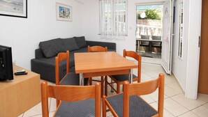Ein gemütlicher Wohnraum mit einer Couch, einem Holzesstisch und Stühlen. Ein Fernseher steht auf einem kleinen Tisch, und ein Fenster mit transparenten Vorhängen ist zu sehen.