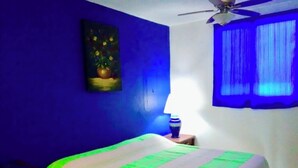 Een kamer met een bed, een plafondventilator, een schilderij aan de muur en een raam met blauwe gordijnen.