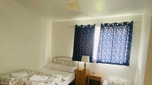 A small, well-lit bedroom with two single beds, a nightstand with a lamp, and a window with star-patterned curtains.