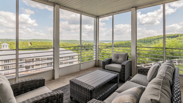 Un balcón iluminado por el sol con muebles de mimbre, amplias ventanas que ofrecen vistas a colinas verdes y edificios.