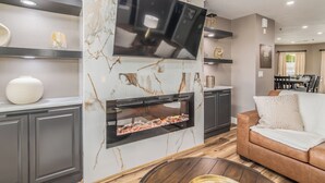 Una sala de estar moderna con un televisor de pantalla plana sobre una chimenea, gabinetes grises, un sofá de cuero marrón y una pared de mármol.