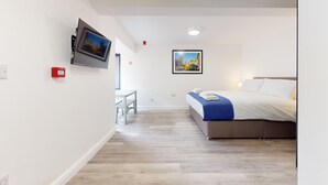 Habitación de hotel con cama, televisor montado en la pared, una mesita y un cuadro en la pared.