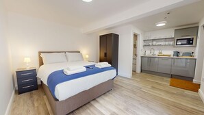 Una habitación de hotel moderna con una cama bien hecha, una mesita de noche pequeña con una lámpara, un armario empotrado y una kitchenette compacta con microondas y fregadero.