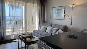 A modern living room with a grey sofa, a dining table, and a view of the sea through sheer curtains.