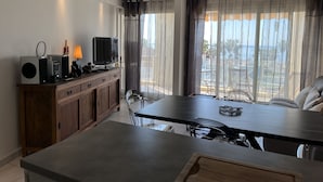 A modern kitchen with a dining area, large windows with curtains, a flat-screen TV, and a wooden cabinet.