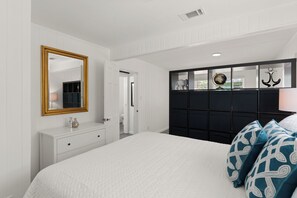 A bedroom with a white bed, a dresser, a mirror, a black headboard with decorative items, and a door leading to another room.