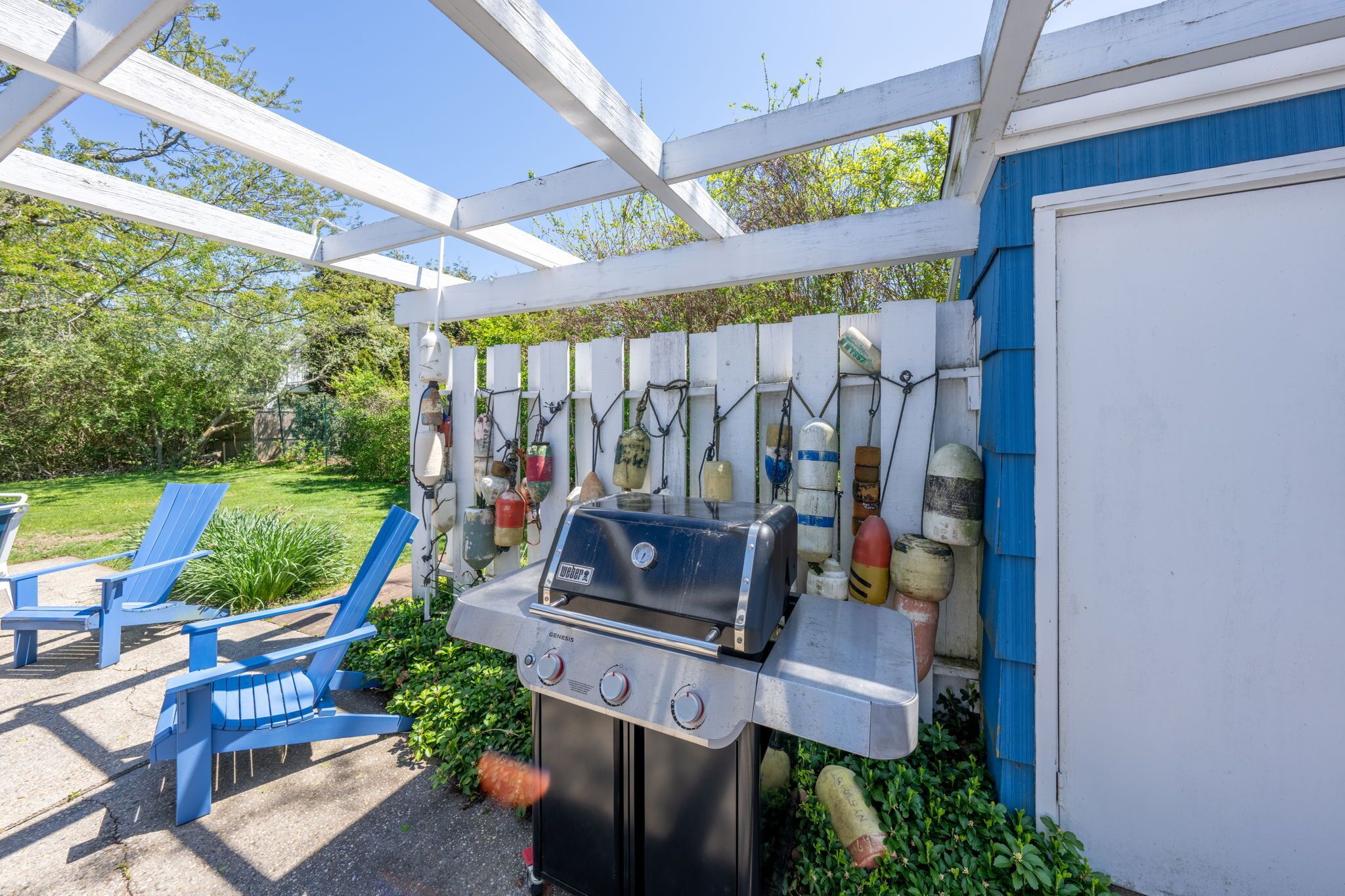 A barbecue grill under a white pergola, surrounded by blue Adirondack chairs and a garden.