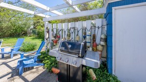 Un barbecue install é sous une pergola blanche, entouré de chaises Adirondack bleues et d’un jardin.