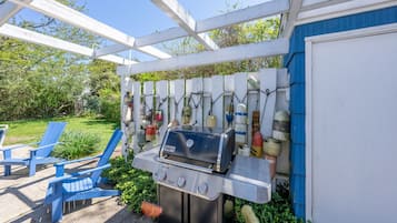 A barbecue grill under a white pergola, surrounded by blue Adirondack chairs and a garden.