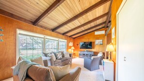 Un salon spacieux avec un plafond haut, des poutres apparentes, une grande fenêtre, un téléviseur à écran plat et un coin salon confortable.