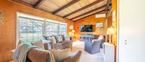 A spacious living room with a high ceiling, wooden beams, a large window, a flat-screen TV, and a cozy seating area.
