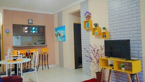 A living room with a TV on a stand, a dining area with chairs, and a kitchen area with a fridge and a window. - Apartment for the season in Praia do Futuro (Antonio Diogo Fortaleza)