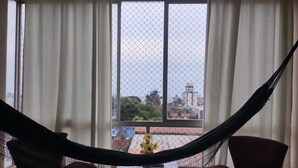 A hammock between two chairs in front of a window with a view of buildings and a yellow flower. - Apartment for the season in Praia do Futuro (Antonio Diogo Fortaleza)