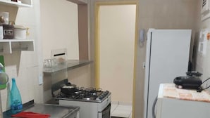 A compact kitchen with a gas stove, oven, and a refrigerator. There is a sink with a red mat, and a shelf with kitchen items. - Apartment for the season in Praia do Futuro (Antonio Diogo Fortaleza)