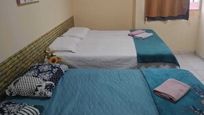 A room with two beds, each with a blue quilted bedspread and white bedding. There is a painting on the wall above the left bed, and a window with a brown curtain on the right. - Apartment for the season in Praia do Futuro (Antonio Diogo Fortaleza)