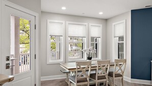 A dining area with a table and chairs, a vase with flowers, and a view through the windows.