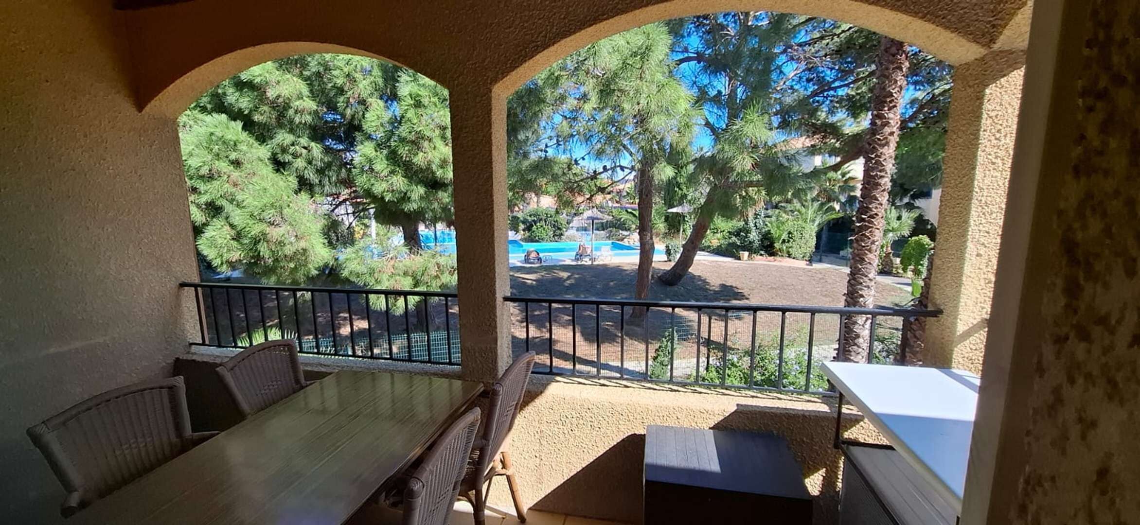 Ein überdachter Außenbereich mit Tisch und Stühlen, mit Blick auf ein Schwimmbad und Bäume.