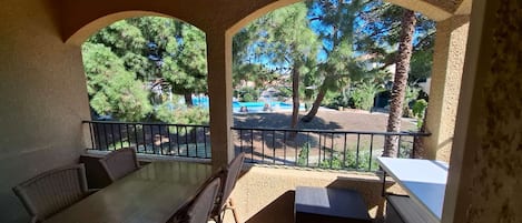 Ett takförsett utomhusområde med bord och stolar, med utsikt över en pool och träd.