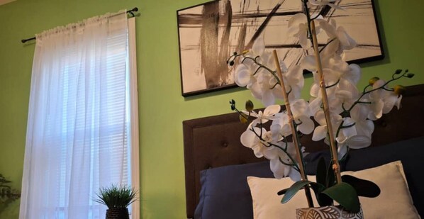 A room with a bed, a window with white curtains, a wall-mounted painting, and a potted orchid plant.