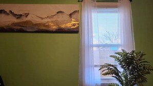 Une pièce avec un mur vert, une fenêtre avec des stores blancs et un tableau représentant un paysage.