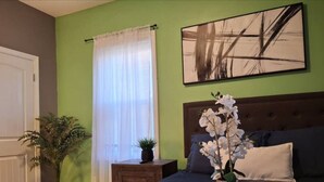 Une chambre à coucher avec un lit, une table de chevet, une plante en pot, une fenêtre avec des rideaux et un tableau au mur.