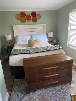 A bedroom with a bed, bedside tables, a dresser, a wall art piece, and a window with blinds.