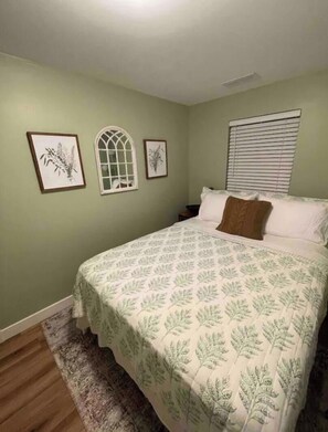 A bedroom with a bed, a nightstand, a window with blinds, and two framed botanical prints on the wall.