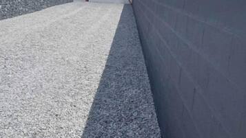 Un bâtiment à l’extérieur blanc et au toit gris, situé à côté d’un mur gris et d’un chemin de gravier.