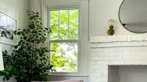 Una habitación acogedora con chimenea de ladrillo blanco, un sillón cómodo con cojín, una mesita y un gran ventanal con cortinas.
