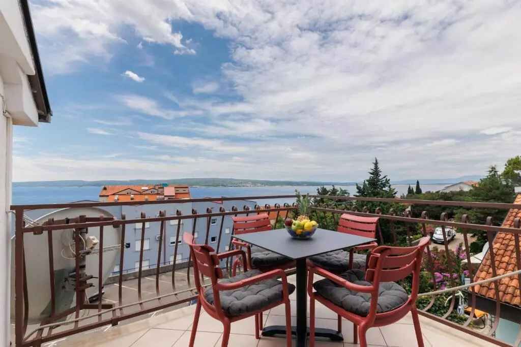 Varanda com mesa e cadeiras, vista para um corpo de água e edifícios ao longe.