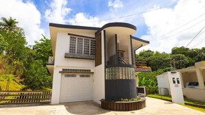 A modern two-story house with a garage, a curved section, and a small balcony. - Villa Campiña Family Home Scenic Mountain Views (Utuado)